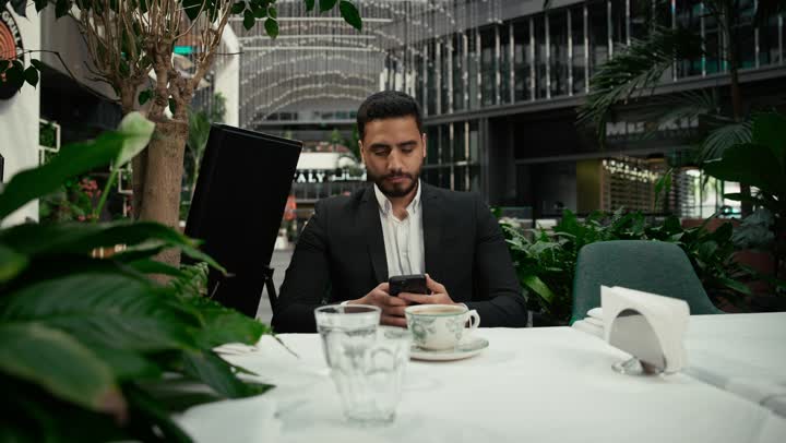 توثيق اللحظات السعيدة، كافيه راقي في الإمارات العربية المتحدة، الاستمتاع بالاجواء الهادئة خارج المنزل، رجل عربي خليجي اماراتي يرتدي البدلة الرسمية يجلس في المقهى يقوم بالتقاط صورة تذكارية لفنجان القهوة باستخدام الهاتف المحمول