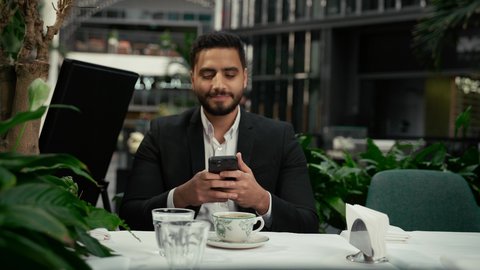 توثيق اللحظات السعيدة، كافيه راقي في الإمارات العربية المتحدة، الاستمتاع بالاجواء الهادئة خارج المنزل، رجل عربي خليجي اماراتي يرتدي البدلة الرسمية يجلس في المقهى يقوم بالتقاط صورة تذكارية لفنجان القهوة باستخدام الهاتف المحمول