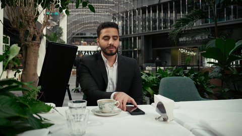 كافيه راقي في الإمارات العربية المتحدة، التواصل مع الأهل والأصدقاء عبر الجوال، الاستمتاع بالاجواء الهادئة خارج المنزل، رجل عربي خليجي اماراتي يرتدي البدلة الرسمية يجلس في المقهى يقوم بشرب القهوة وإجراء مكالمة هاتفية باستخدام الهاتف المحمول بتعابير السرور