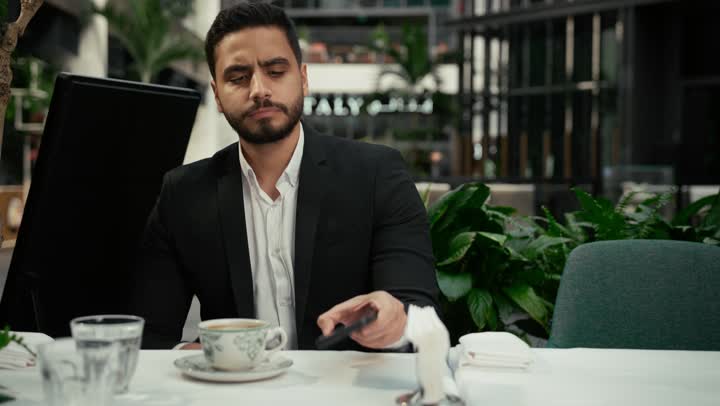 كافيه راقي في الإمارات العربية المتحدة، التواصل مع الأهل والأصدقاء عبر الجوال، الاستمتاع بالاجواء الهادئة خارج المنزل، رجل عربي خليجي اماراتي يرتدي البدلة الرسمية يجلس في المقهى يقوم بشرب القهوة وإجراء مكالمة هاتفية باستخدام الهاتف المحمول بتعابير السرور