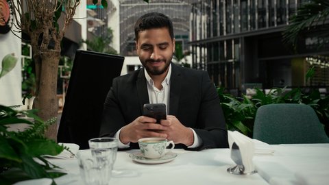 تعابير التلذذ والاستمتاع باحتساء القهوة، تصفح مواقع التواصل الاجتماعي عبر الجوال، الاستمتاع بالاجواء الهادئة خارج المنزل، تصوير مقرب لرجل عربي خليجي اماراتي يرتدي زي رسمي يجلس في المقهى يقوم باستخدام الهاتف المحمول، كافيه راقي في الإمارات العربية المتحدة