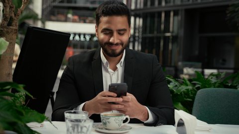 تعابير التلذذ والاستمتاع باحتساء القهوة، تصفح مواقع التواصل الاجتماعي عبر الجوال، الاستمتاع بالاجواء الهادئة خارج المنزل، تصوير مقرب لرجل عربي خليجي اماراتي يرتدي زي رسمي يجلس في المقهى يقوم باستخدام الهاتف المحمول، كافيه راقي في الإمارات العربية المتحدة