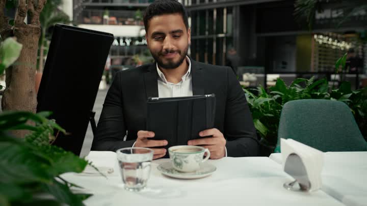 إنجاز العمل عن بعد عبر التابلت، التلذذ والاستمتاع باحتساء القهوة، شاب عربي خليجي إماراتي مبتسم يرتدي الزي الرسمي يجلس في المقهى ويقوم باستخدام الجهاز اللوحي المحمول بإيماءات الاندماج، جلسة خارجية هادئة في كافيه راقي في الإمارات، استخدام جهاز تقني حديث ومتطور