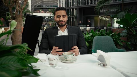 إنجاز العمل عن بعد عبر التابلت، التلذذ والاستمتاع باحتساء القهوة، شاب عربي خليجي إماراتي مبتسم يرتدي الزي الرسمي يجلس في المقهى ويقوم باستخدام الجهاز اللوحي المحمول بإيماءات الاندماج، جلسة خارجية هادئة في كافيه راقي في الإمارات، استخدام جهاز تقني حديث ومتطور