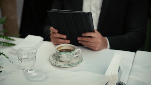 جلسة خارجية هادئة في كافيه راقي في الإمارات، استخدام جهاز تقني حديث ومتطور، إنجاز العمل عن بعد عبر التابلت، التلذذ والاستمتاع باحتساء القهوة، تصوير مقرب لأيدي شاب عربي خليجي إماراتي يقوم باستخدام الجهاز اللوحي المحمول