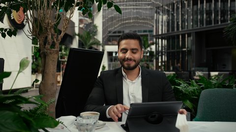 رفع اليد بإيماءات إلقاء التحية والسلام، التلذذ والاستمتاع باحتساء القهوة، تصوير مقرب لرجل عربي خليجي إماراتي يرتدي الزي الرسمي يجلس في المقهى ويقوم بإنجاز العمل عن بعد عبر جهاز التابلت، جلسة خارجية هادئة في كافيه راقي في الإمارات، استخدام جهاز تقني حديث ومتطور