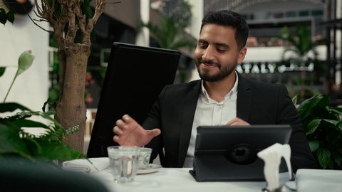 رفع اليد بإيماءات إلقاء التحية والسلام، التلذذ والاستمتاع باحتساء القهوة، تصوير مقرب لرجل عربي خليجي إماراتي يرتدي الزي الرسمي يجلس في المقهى ويقوم بإنجاز العمل عن بعد عبر جهاز التابلت، جلسة خارجية هادئة في كافيه راقي في الإمارات، استخدام جهاز تقني حديث ومتطور