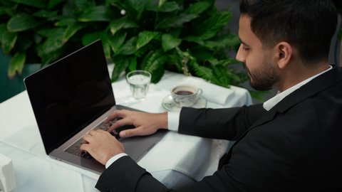 عرض شاشة لابتوب سوداء فارغة، امتلاك مهارة الكتابة بسرعة، الاستمتاع باحتساء القهوة، تصوير مقرب من الجانب لرجل عربي خليجي إماراتي يرتدي الزي الرسمي يجلس في المقهى ويقوم بالضغط على أزار لوحة المفاتيح، إنجاز العمل عن بعد عبر جهاز الحاسوب، كافيه راقي في الإمارات