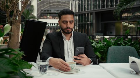 توثيق اللحظات السعيدة، الاستمتاع بالاجواء الهادئة خارج المنزل، رجل عربي خليجي اماراتي يرتدي البدلة الرسمية يجلس في إحدى المقاهي الراقية يقوم بالتقاط صورة سيلفي تذكارية باستخدام الهاتف المحمول بإيماءات السرور، التلذذ باحتساء فنجان من القهوة الساخنة