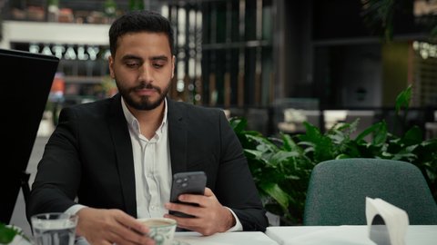 توثيق اللحظات السعيدة، الاستمتاع بالاجواء الهادئة خارج المنزل، رجل عربي خليجي اماراتي يرتدي البدلة الرسمية يجلس في إحدى المقاهي الراقية يقوم بالتقاط صورة سيلفي تذكارية باستخدام الهاتف المحمول بإيماءات السرور، التلذذ باحتساء فنجان من القهوة الساخنة