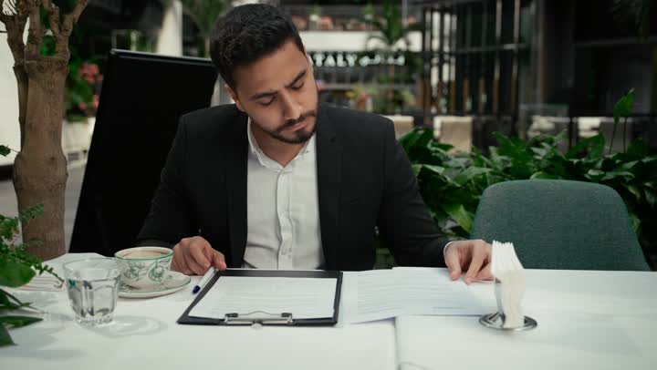بناء شراكات واتفاقيات جديدة، احتساء القهوة الساخنة في الكافيه، الموافقة على العرض المالي، قراءة البنود الخاصة بالعمل، التحقق والتدقيق في شروط العقد، تصوير مقرب لرجل عربي خليجي اماراتي يرتدي بدلة رسمية يجلس في إحدى المقاهي ويقوم بالتوقيع على العقود