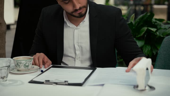 بناء شراكات واتفاقيات جديدة، احتساء القهوة الساخنة في الكافيه، الموافقة على العرض المالي، قراءة البنود الخاصة بالعمل، التحقق والتدقيق في شروط العقد، تصوير مقرب لرجل عربي خليجي اماراتي يرتدي بدلة رسمية يجلس في إحدى المقاهي ويقوم بالتوقيع على العقود