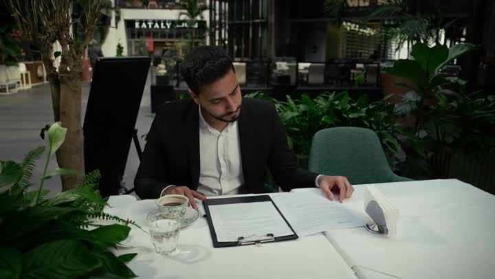 بناء شراكات واتفاقيات جديدة، احتساء القهوة الساخنة في الكافيه، الموافقة على العرض المالي، قراءة البنود الخاصة بالعمل، التحقق والتدقيق في شروط العقد، تصوير مقرب لرجل عربي خليجي اماراتي يرتدي بدلة رسمية يجلس في إحدى المقاهي ويقوم بالتوقيع على العقود