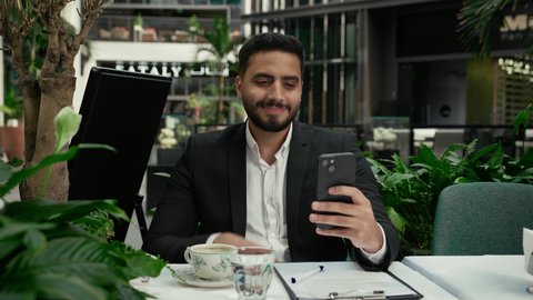 التلذذ باحتساء القهوة الساخنة، مشاركة الأخبار والأحاديث الجيدة، رفع اليد بإيماءات إلقاء التحية والسلام، الاستمتاع بالاجواء الهادئة خارج المنزل، رجل عربي خليجي إماراتي يرتدي البدلة الرسمية يجلس في المقهى ويقوم بإجراء مكالمة فيديو عبر الهاتف الجوال بتعابير السرور