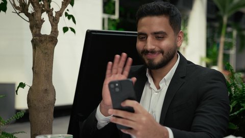 مشاركة الأخبار والأحاديث الجيدة، رفع اليد بإيماءات إلقاء التحية والسلام، الاستمتاع بالاجواء الهادئة خارج المنزل، تصوير مقرب لرجل عربي خليجي إماراتي يرتدي البدلة الرسمية يجلس في المقهى ويقوم بإجراء مكالمة فيديو عبر الهاتف الجوال بتعابير السرور