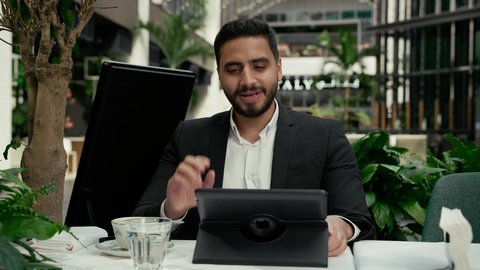 رفع اليد بإيماءات إلقاء التحية والسلام، الاستمتاع بالاجواء الهادئة خارج المنزل، رجل عربي خليجي إماراتي يرتدي البدلة الرسمية يجلس في المقهى ويقوم بإجراء مكالمة فيديو عبر جهاز التابلت بتعابير السرور، مشاركة الأخبار والأحاديث الجيدة، عقد اجتماع عمل عن بعد