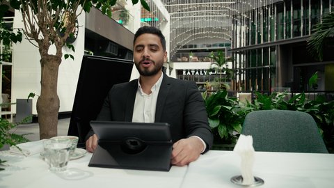رفع اليد بإيماءات إلقاء التحية والسلام، الاستمتاع بالاجواء الهادئة خارج المنزل، رجل عربي خليجي إماراتي يرتدي البدلة الرسمية يجلس في المقهى ويقوم بإجراء مكالمة فيديو عبر جهاز التابلت بتعابير السرور، مشاركة الأخبار والأحاديث الجيدة، عقد اجتماع عمل عن بعد