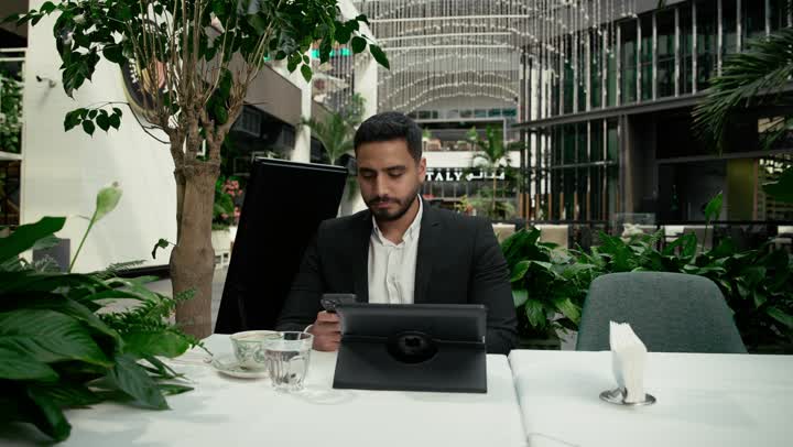 إيماءات الصدمة والغضب بتلقي الأخبار السيئة، جلسة خارجية هادئة في كافيه راقي في الإمارات العربية المتحدة, تصوير مقرب لرجل عربي خليجي إماراتي يرتدي البدلة الرسمية يجلس في المقهى ويقوم بإنجاز العمل عن بعد عبر جهاز التابلت، تصفح مواقع الانترنت عبر الهاتف المحمول