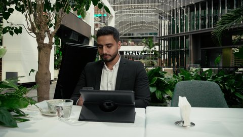 جلسة خارجية هادئة في كافيه راقي في الإمارات العربية المتحدة, تصوير مقرب لرجل عربي خليجي إماراتي يرتدي البدلة الرسمية يجلس في المقهى ويقوم بإنجاز العمل عن بعد عبر جهاز التابلت، إيماءات الصدمة والغضب بتلقي الأخبار السيئة