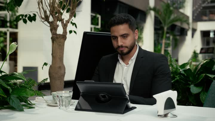 جلسة خارجية هادئة في كافيه راقي في الإمارات العربية المتحدة, تصوير مقرب لرجل عربي خليجي إماراتي يرتدي البدلة الرسمية يجلس في المقهى ويقوم بإنجاز العمل عن بعد عبر جهاز التابلت، إيماءات الصدمة والغضب بتلقي الأخبار السيئة