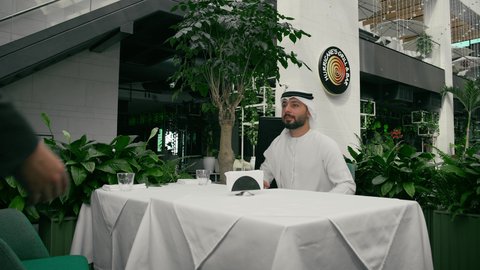 مفهوم الاستقبال والترحيب، إيماءات السعادة والسرور برؤية الأصدقاء، إلقاء التحيه والسلام, صداقة وتعارف وبناء علاقات اجتماعية، رجل عربي خليجي إماراتي يرتدي الكندورة والغترة يقابل صديقه في المقهى ويقوم بمصافحته، كافيه راقي في الإمارات العربية المتحدة