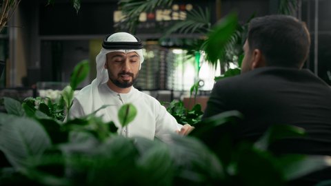 تبادل الاحاديث بين الأصدقاء، قضاء وقت ممتع في الخارج، مشاركة المشروبات الساخنة والقهوة، رجل عربي خليجي اماراتي يرتدي الكندورة والغترة البيضاء يجلس مع صديق يرتدي البدلة الرسمية في المقهى، جلسة تعارف وصداقة في إحدى الكافيهات الراقية في الإمارات العربية المتحدة