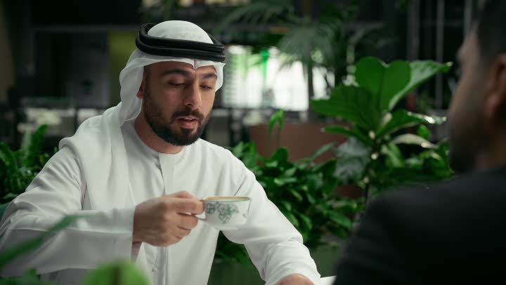 تبادل الاحاديث بين الأصدقاء، قضاء وقت ممتع في الخارج، مشاركة المشروبات الساخنة والقهوة، رجل عربي خليجي اماراتي يرتدي الكندورة والغترة البيضاء يجلس مع صديق يرتدي البدلة الرسمية في المقهى، جلسة تعارف وصداقة في إحدى الكافيهات الراقية في الإمارات العربية المتحدة