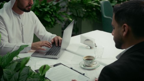 انجاز المهام عن بعد عبر الحاسوب المحمول في الكافيه، عقد صفقات عمل جديدة، الاستمتاع بشرب القهوة في المقهى، استخدام التقنيات الحديثة والمطورة، تصوير مقرب لرجل عربي خليجي اماراتي يرتدي البدلة الرسمية في اجتماع عمل مع عميل يرتدي الزي التقليدي.