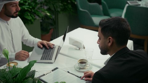 انجاز المهام عن بعد عبر الحاسوب المحمول في الكافيه، عقد صفقات عمل جديدة، الاستمتاع بشرب القهوة في المقهى، استخدام التقنيات الحديثة والمطورة، تصوير مقرب لرجل عربي خليجي اماراتي يرتدي البدلة الرسمية في اجتماع عمل مع عميل يرتدي الزي التقليدي.
