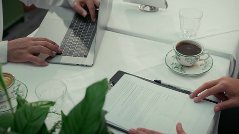 إنجاز العمل عن بعد باستخدام الحاسوب المحمول، قراءة البنود الخاصة بالعمل، التحقق والتدقيق في شروط العقد، تصوير مقرب لأيدي رجلين عربيين خليجيين اماراتيين يجلسان في إحدى المقاهي يقومان بقراءة أوراق العقود والتقارير، بناء شراكات واتفاقيات جديدة، احتساء القهوة الساخنة