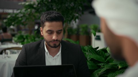 الاطلاع والموافقة على التقارير والمشاريع الجديدة، انجاز المهام عن بعد عبر الحاسوب المحمول، تصوير مقرب من الخلف لرجل عربي خليجي اماراتي يرتدي الزي التقليدي يجلس في المقهى في اجتماع عمل مع عميل يرتدي البدلة الرسمية، استخدام التقنيات الحديثة والمطورة.
