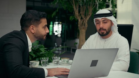 إيماءات الجدية والتركيز، الحوار والنقاش بين الزملاء لإدارة الأعمال وتنظيمها، استخدام التقنيات الحديثة والمطورة، تصوير مقرب لشابين عربيين خليجيين إماراتيين يجلسان في المقهى لإنجاز المشاريع ومهام الأعمال عن بعد باستخدام جهاز الحاسوب المحمول، تناول القهوة الساخنة