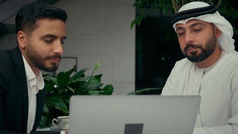 إيماءات الجدية والتركيز، الحوار والنقاش بين الزملاء لإدارة الأعمال وتنظيمها، استخدام التقنيات الحديثة والمطورة، تصوير مقرب لشابين عربيين خليجيين إماراتيين يجلسان في المقهى لإنجاز المشاريع ومهام الأعمال عن بعد باستخدام جهاز الحاسوب المحمول، تناول القهوة الساخنة