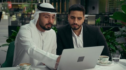 إيماءات الجدية والتركيز، الحوار والنقاش بين الزملاء لإدارة الأعمال وتنظيمها، استخدام التقنيات الحديثة والمطورة، تصوير مقرب لشابين عربيين خليجيين إماراتيين يجلسان في المقهى لإنجاز المشاريع ومهام الأعمال عن بعد باستخدام جهاز الحاسوب المحمول، تناول القهوة الساخنة