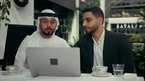 إيماءات الجدية والتركيز، الحوار والنقاش بين الزملاء لإدارة الأعمال وتنظيمها، استخدام التقنيات الحديثة والمطورة، تصوير مقرب لشابين عربيين خليجيين إماراتيين يجلسان في المقهى لإنجاز المشاريع ومهام الأعمال عن بعد باستخدام جهاز الحاسوب المحمول، تناول القهوة الساخنة