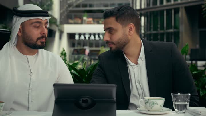 الحوار والنقاش بين الزملاء لإدارة الأعمال وتنظيمها، استخدام التقنيات الحديثة والمطورة في العمل، انجاز المشاريع ومهام الأعمال عن بعد في الكافيه، رجل عربي خليجي اماراتي في اجتماع عمل مع عميل جديد يرتدي البدلة الرسمية، استخدام جهاز التابلت المحمول