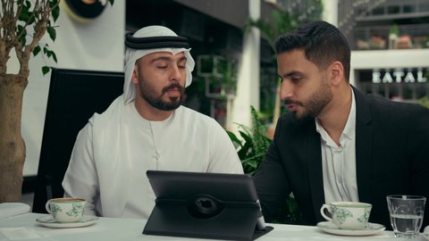 الحوار والنقاش بين الزملاء لإدارة الأعمال وتنظيمها، استخدام التقنيات الحديثة والمطورة في العمل، انجاز المشاريع ومهام الأعمال عن بعد في الكافيه، رجل عربي خليجي اماراتي في اجتماع عمل مع عميل جديد يرتدي البدلة الرسمية، استخدام جهاز التابلت المحمول