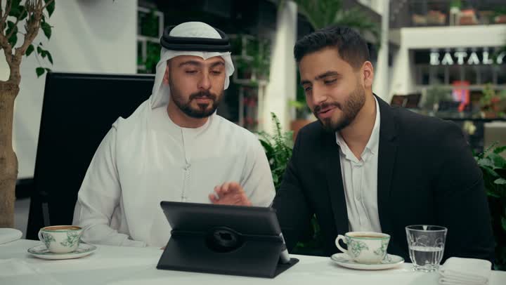 الحوار والنقاش بين الزملاء لإدارة الأعمال وتنظيمها، استخدام التقنيات الحديثة والمطورة في العمل، انجاز المشاريع ومهام الأعمال عن بعد في الكافيه، تصوير مقرب لزميلين عربيين خليجيين إماراتيين يجلسان في المقهى يقومان بإجراء اجتماع عمل عن بعد عبر جهاز التابلت المحمول