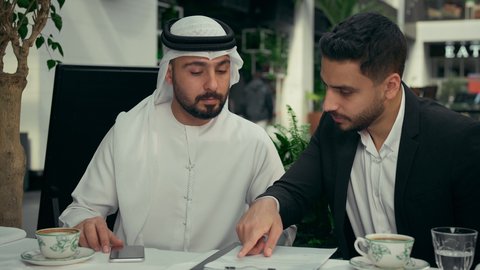 الرد علة مكالمة هاتفية عبر الهاتف الجوال، الحوار والنقاش بين الزملاء لإدارة الأعمال وتنظيمها، استخدام التقنيات الحديثة والمطورة، انجاز المشاريع ومهام الأعمال في الكافيه، تصوير مقرب لزميلين عربيين خليجيين إماراتيين يجلسان في المقهى يقومان بإجراء اجتماع عمل