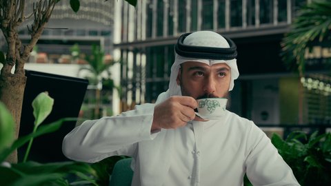 التلذذ باحتساء مشروبات الكافيهات الجاهزة، كافيه راقي في الإمارات العربية المتحدة، تأثير الكافيين على نشاط الدماغ والذاكرة، الاستمتاع بالاجواء الهادئة خارج المنزل، تصوير مقرب لرجل عربي خليجي اماراتي يرتدي الكندورة والغترة يجلس في المقهى يقوم بشرب القهوة