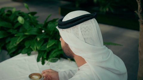 التلذذ باحتساء مشروبات الكافيهات الجاهزة، كافيه راقي في الإمارات العربية المتحدة، تأثير الكافيين على نشاط الدماغ والذاكرة، الاستمتاع بالاجواء الهادئة خارج المنزل، تصوير مقرب من الجانب لرجل عربي خليجي اماراتي يرتدي الكندورة والغترة يجلس في المقهى يقوم بشرب القهوة