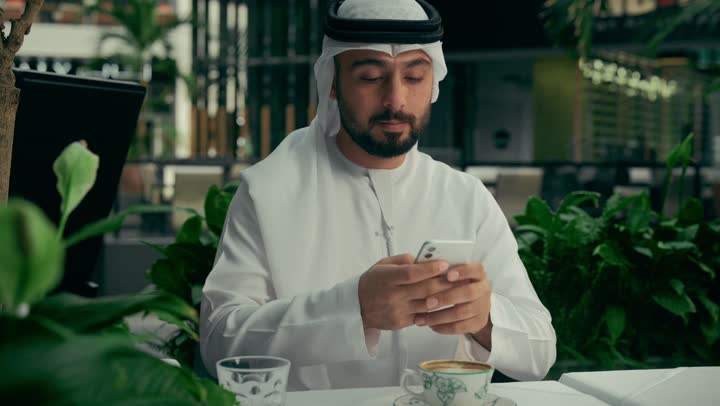 تصفح مواقع التواصل الاجتماعي عبر الجوال، الاستمتاع بالاجواء الهادئة خارج المنزل، تصوير مقرب لرجل عربي خليجي اماراتي يرتدي الكندورة والغترة يجلس في المقهى يقوم بشرب القهوة واستخدام الهاتف المحمول، كافيه راقي في الإمارات العربية المتحدة