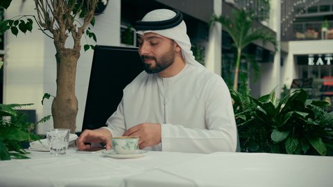 كافيه راقي في الإمارات العربية المتحدة، التواصل مع الأهل والأصدقاء عبر الجوال، الاستمتاع بالاجواء الهادئة خارج المنزل، رجل عربي خليجي اماراتي يرتدي الكندورة والغترة يجلس في المقهى يقوم بشرب القهوة وإجراء مكالمة هاتفية باستخدام الهاتف المحمول