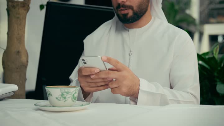 كافيه راقي في الإمارات العربية المتحدة، تصفح مواقع التواصل الاجتماعي عبر الجوال، الاستمتاع بالاجواء الهادئة خارج المنزل، تصوير مقرب لرجل عربي خليجي اماراتي يرتدي الكندورة والغترة يجلس في المقهى يقوم بشرب القهوة واستخدام الهاتف المحمول