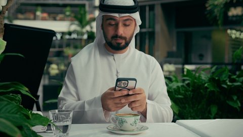 تصفح مواقع التواصل الاجتماعي عبر الجوال، الاستمتاع بالاجواء الهادئة خارج المنزل، تصوير مقرب لرجل عربي خليجي اماراتي يرتدي الكندورة والغترة يجلس في المقهى يقوم بشرب القهوة واستخدام الهاتف المحمول، كافيه راقي في الإمارات العربية المتحدة