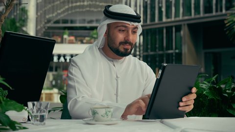 الاستمتاع بالاجواء الهادئة خارج المنزل، رجل عربي خليجي اماراتي يرتدي الكندورة والغترة البيضاء يجلس في إحدى المقاهي الراقية يقوم بتناول فنجان من القهوة واستخدام جهاز التابلت، إنجاز مهام العمل عن بعد عبر الجهاز اللوحي، تصفح مواقع الانترنت