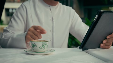 إنجاز مهام العمل عن بعد عبر الجهاز اللوحي، الاستمتاع بالاجواء الهادئة خارج المنزل، تصوير مقرب لرجل عربي خليجي اماراتي يرتدي الكندورة والغترة البيضاء يجلس في إحدى المقاهي الراقية يقوم بتناول فنجان من القهوة واستخدام جهاز التابلت، تصفح مواقع الانترنت