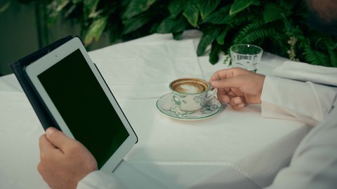 مشاهدة الافلام والمسلسلات المفضلة، إنجاز مهام العمل عن بعد عبر الجهاز اللوحي، الاستمتاع بالاجواء الهادئة خارج المنزل، تصوير مقرب لرجل عربي خليجي اماراتي يجلس في إحدى المقاهي الراقية يقوم بتناول فنجان من القهوة واستخدام التابلت، عرض شاشة سوداء فارغة
