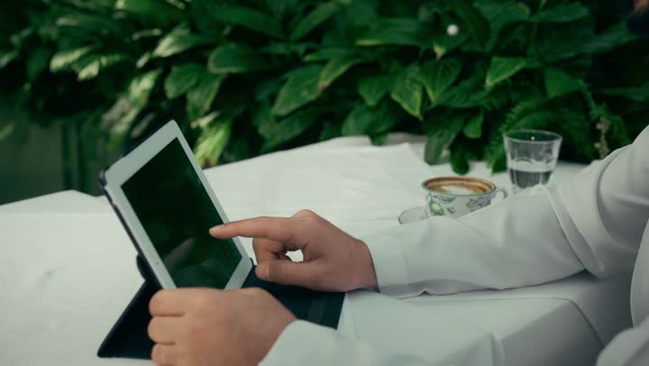 مشاهدة الافلام والمسلسلات المفضلة، إنجاز مهام العمل عن بعد عبر الجهاز اللوحي، الاستمتاع بالاجواء الهادئة خارج المنزل، تصوير مقرب لرجل عربي خليجي اماراتي يجلس في إحدى المقاهي الراقية يقوم بتناول فنجان من القهوة واستخدام التابلت، عرض شاشة سوداء فارغة