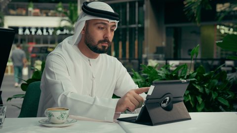 الاستمتاع بالاجواء الهادئة خارج المنزل، رجل عربي خليجي اماراتي يرتدي الكندورة والغترة البيضاء يجلس في إحدى المقاهي الراقية يقوم بتناول فنجان من القهوة واستخدام جهاز التابلت، إنجاز مهام العمل عن بعد عبر الجهاز اللوحي، تصفح مواقع الانترنت